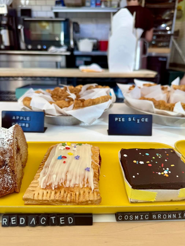 A yellow tray holds a frosted pastry with colorful sprinkles and a chocolate brownie with sprinkles, labeled “REDACTED” and “COSMIC BROWNIE.”